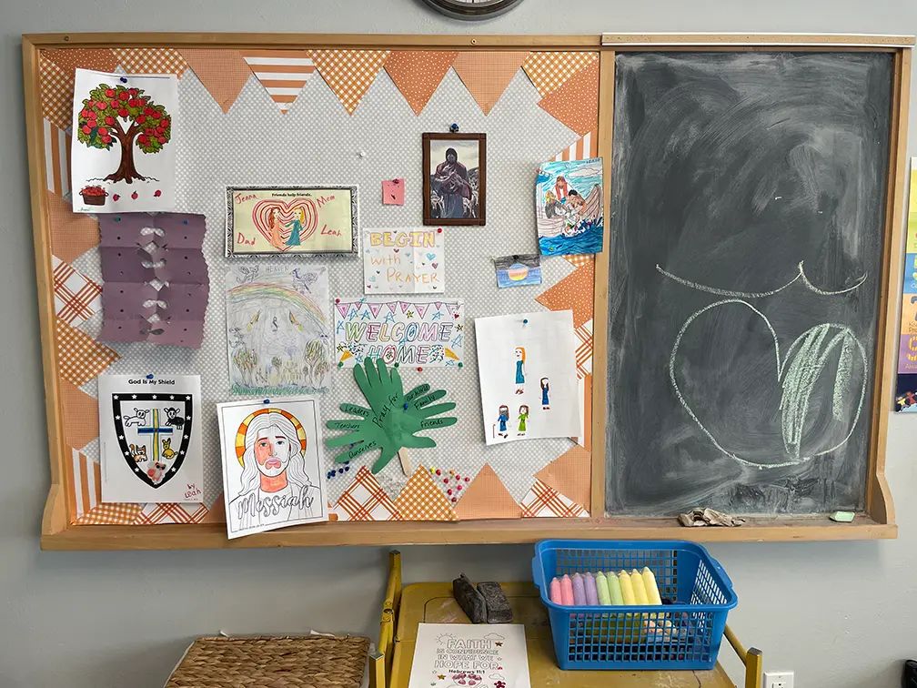A Children’s Church classroom wall with a bulletin board of children’s drawings and a Welcome Home sign next to a chalkboard.