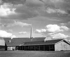 The completed church around 1963, a long low building with a tall spire rising from the roof under a wide cloudy sky