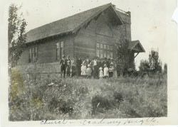 People gathered in front of the first church, a wooden building with a steep roof, on its dedication day in 1920