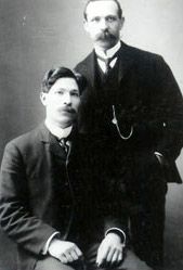 Fridolf Nelson and Fingal Gothberg in dark suits, one seated and the other standing behind him