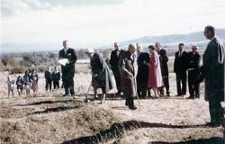 A group of adults in dress clothes gathered on bare ground at the building site for the groundbreaking, with a few children watching from the left