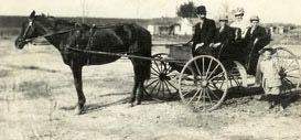 A horse hitched to an open buggy carrying four people in dark coats and hats, with a child standing beside it