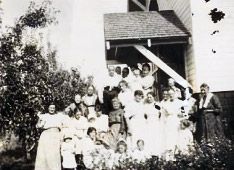 Members of the Ladies Aid Society, mostly women in white dresses, posed outside a building with a covered porch