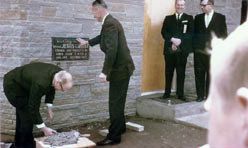 Men in suits setting the cornerstone into a stone wall, one bending over the mortar while others look on