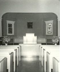 The old church sanctuary, with rows of pews leading to a simple pulpit and communion table, flower arrangements, and framed panels on the wall