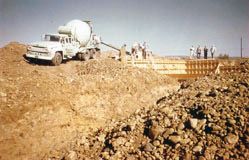 A cement truck pouring concrete into the foundation forms at the building site, with workers standing nearby